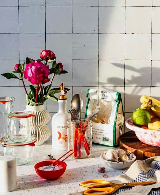 Orange silver cutlery on a counter with fruits, bowls, food, containers, flowers in a kitchen